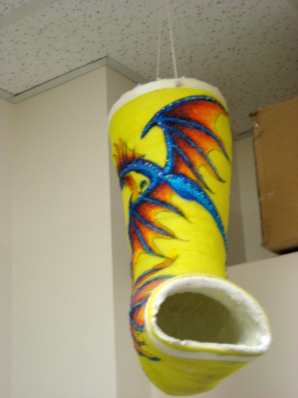 A glittery blue dragon with orange wings against a bright yellow fiberglass foot cast.